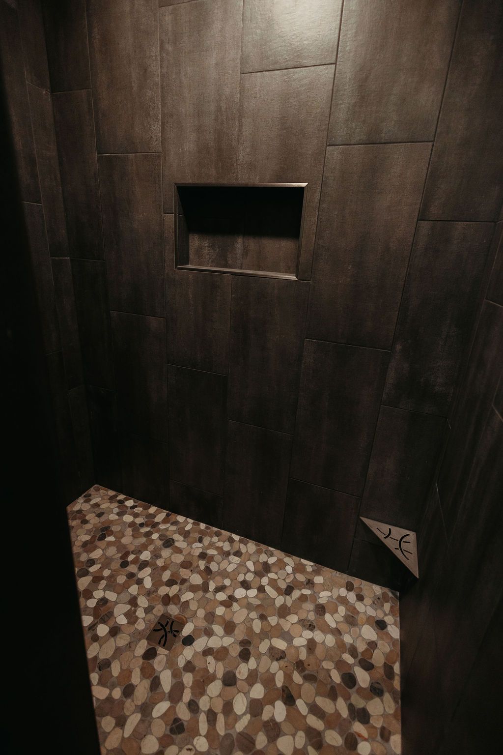 Shower interior with dark brown tile walls, pebble floor, and recessed shelf.
