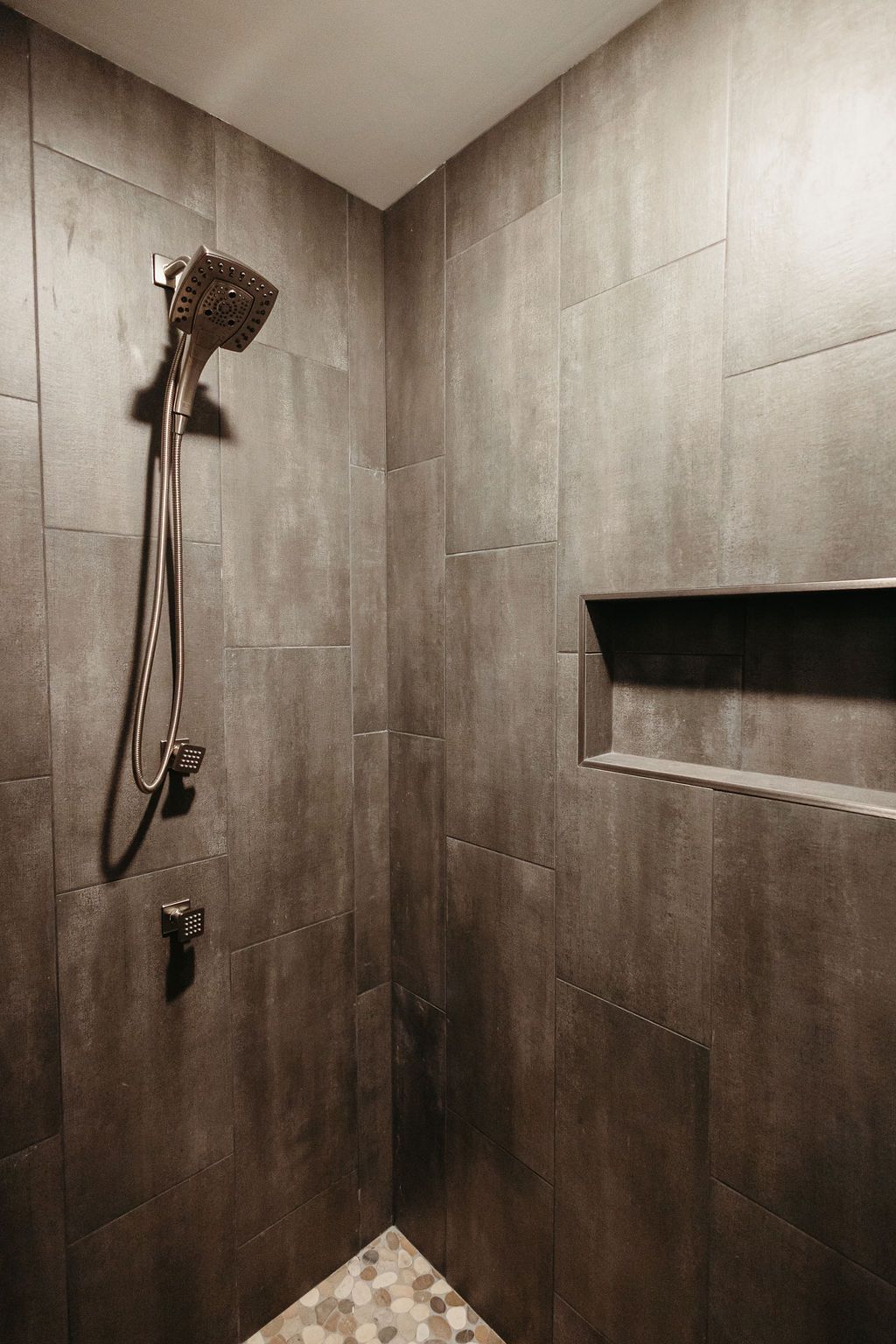 Dark gray tiled shower with built-in shelf, showerhead, and pebble floor corner.