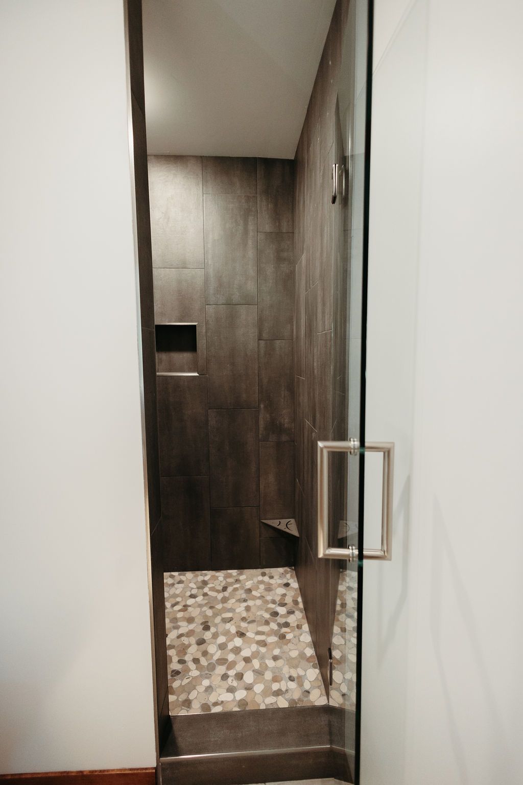 A narrow, modern shower with a glass door, pebble floor, and dark brown tiled walls.