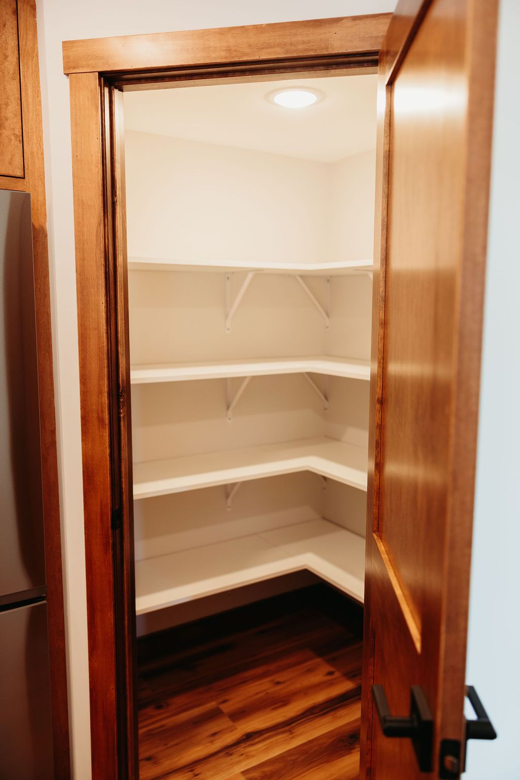 A pantry with white shelves is visible through a partially open wooden door. The walls are white and the floor is wood.