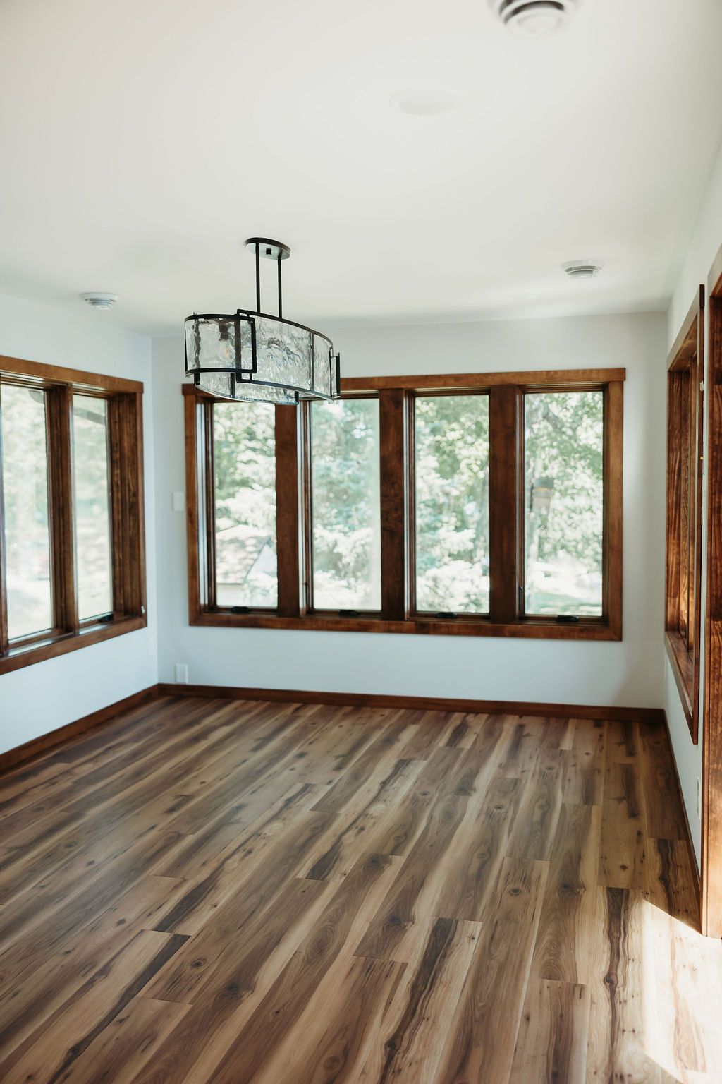 Empty room with hardwood floors, dark wood-framed windows, white ceiling, and a modern chandelier.