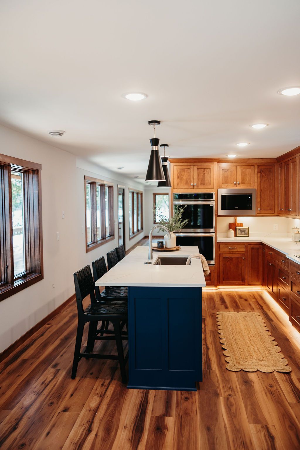 Kitchen with blue island, wood cabinets, and pendant lights. Wooden floor and long row of windows.