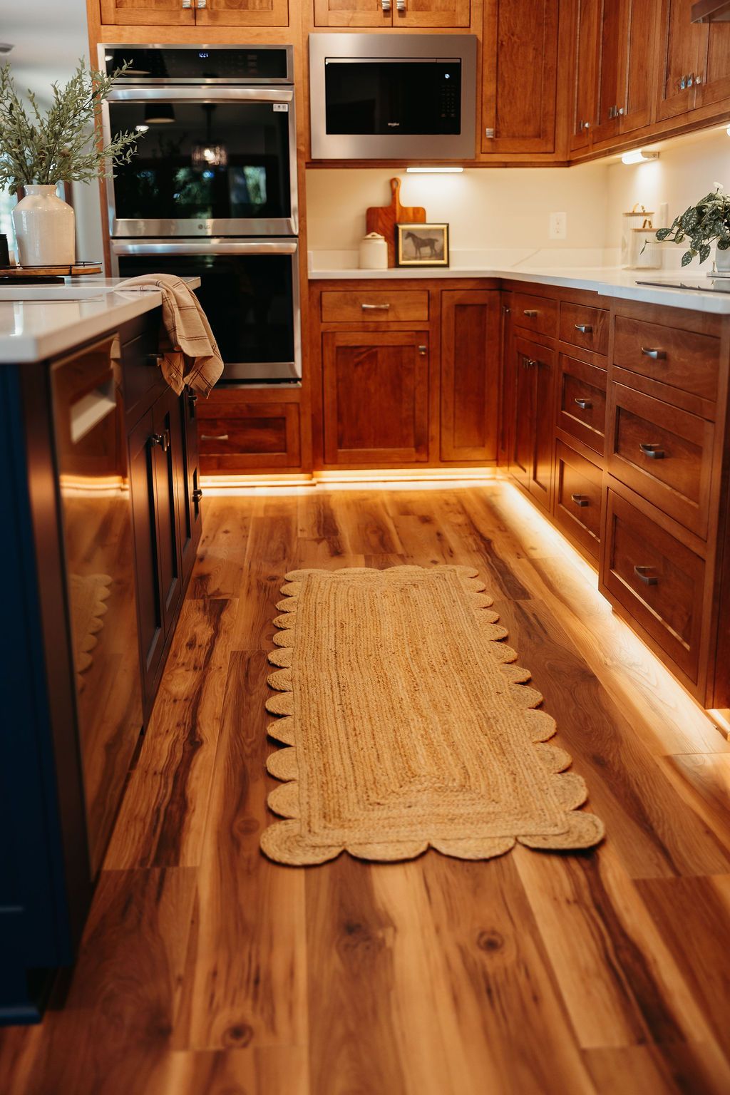 Warm-toned kitchen with wooden cabinets, a blue island, and under-cabinet lighting. A woven rug sits on the wood floor.