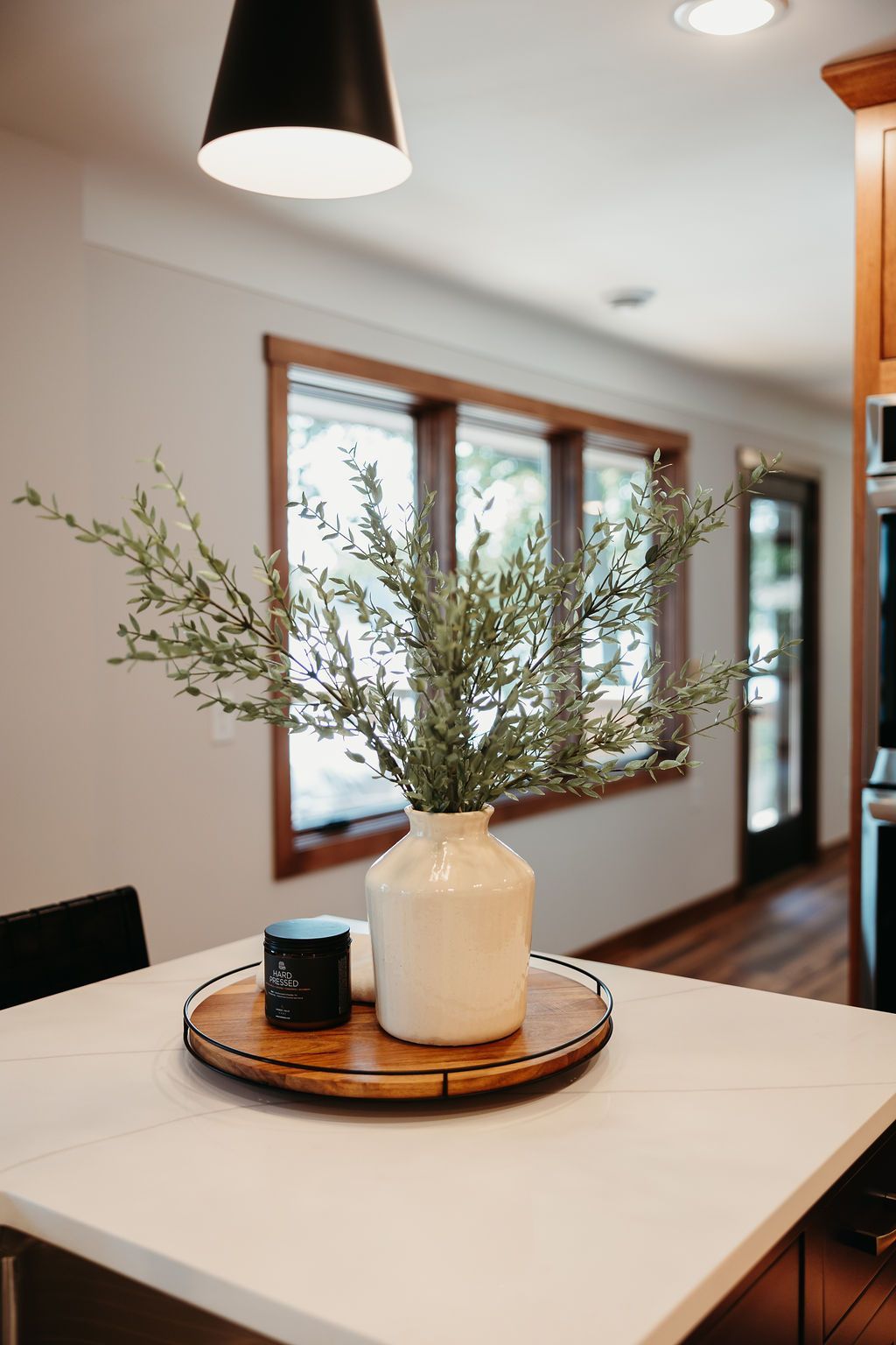 A white vase with greenery on a wooden tray atop a kitchen island. A window and hanging light are visible.