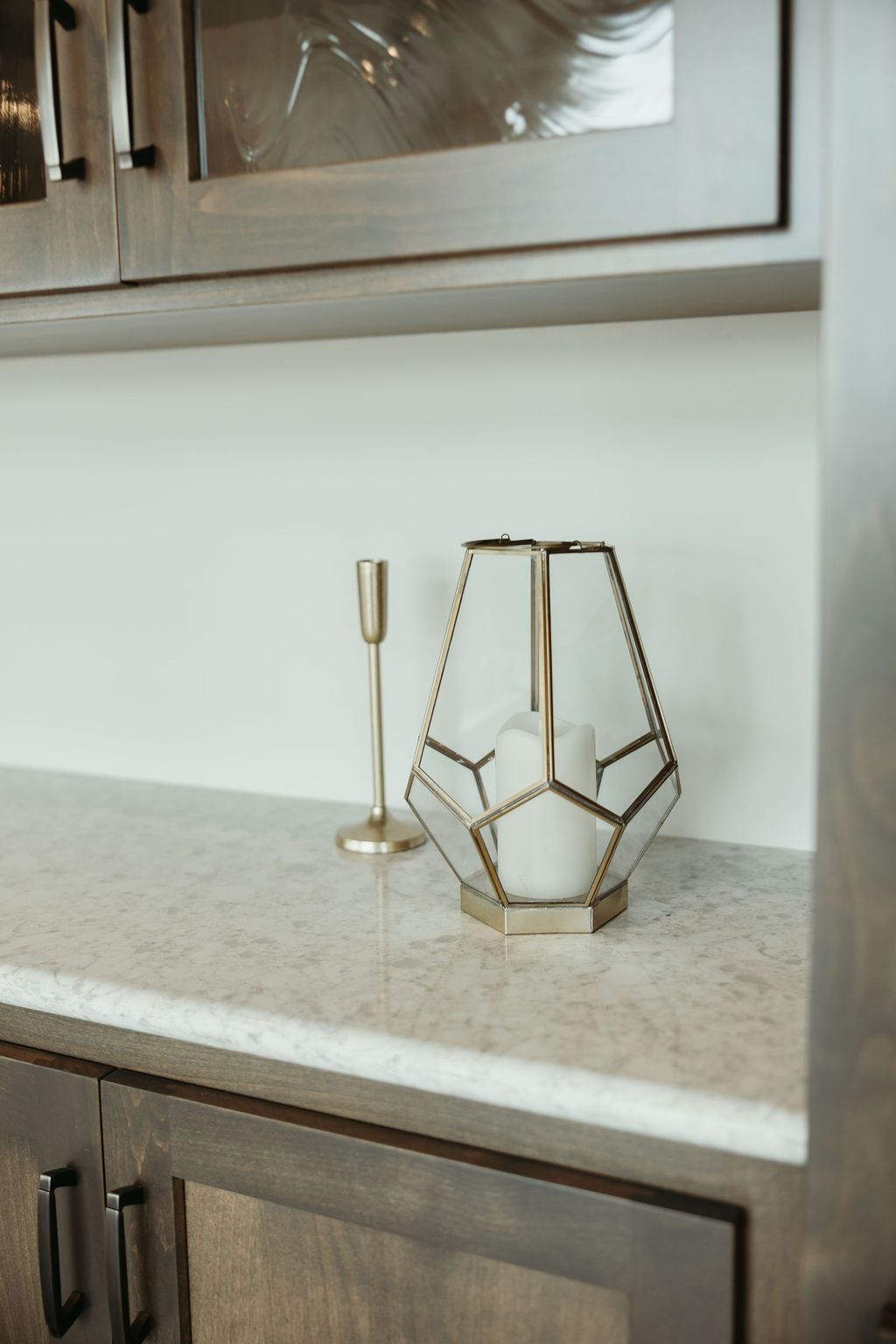 White and gold geometric candle holder and gold candlestick on a countertop. Cabinets above and below.