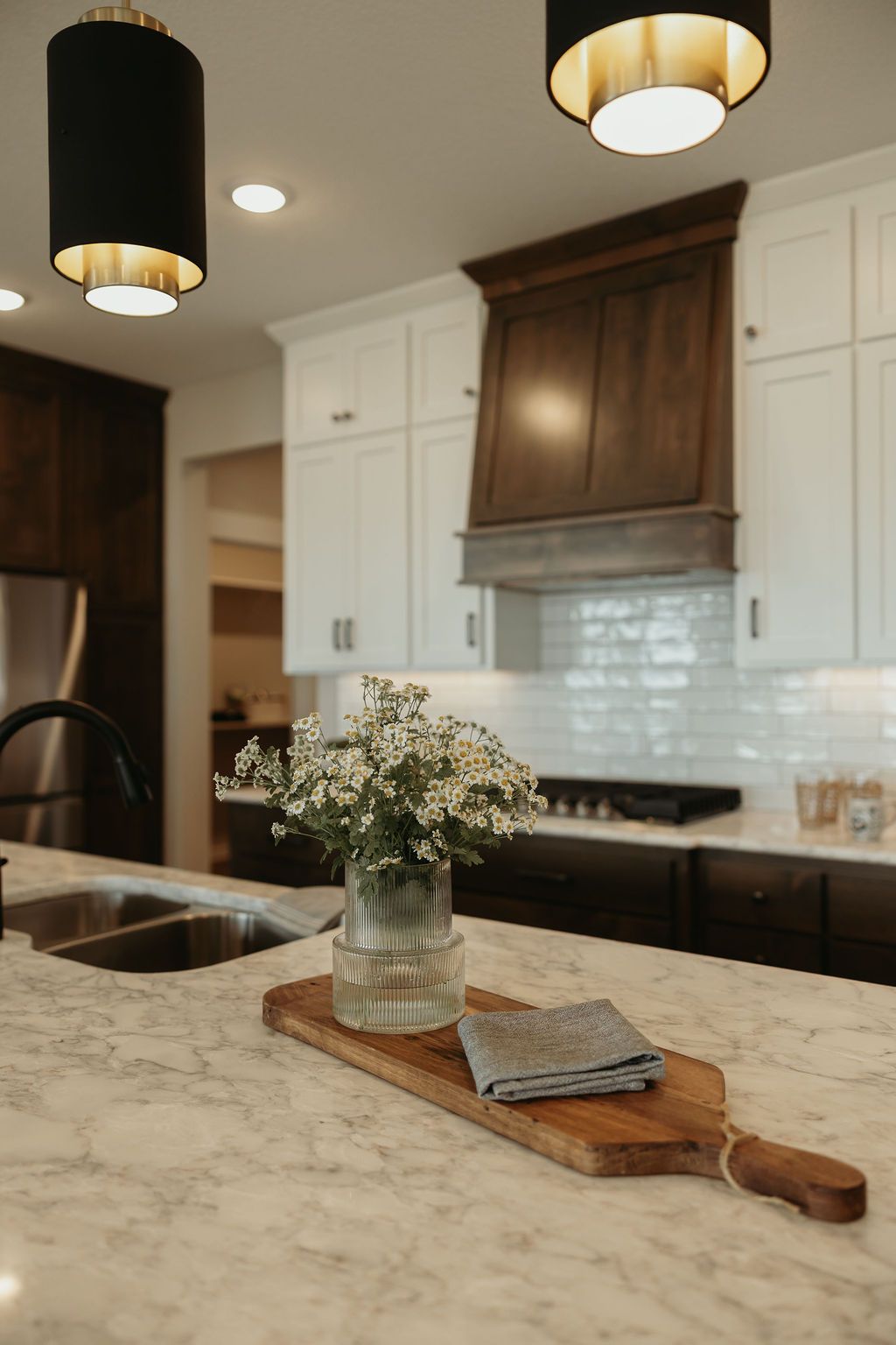 Kitchen island with flowers, a wooden board, and overhead lighting. White cabinets, dark range hood.