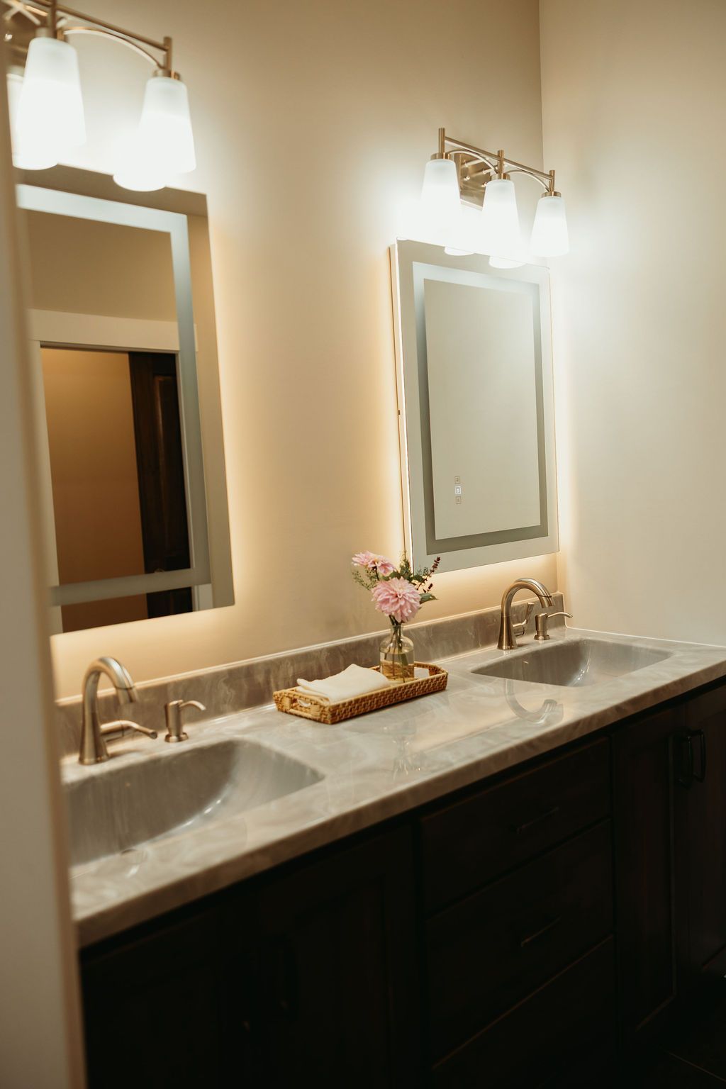 Bathroom with two sinks, mirrors, and sconce lighting above a marble countertop. Dark cabinets. Pink flowers.
