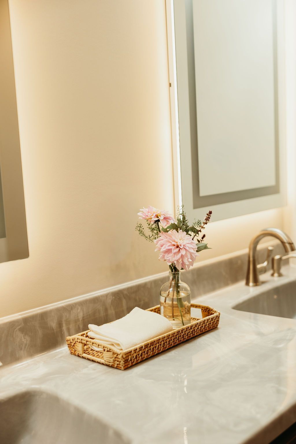 Bathroom sink with pink flowers in vase, stacked white washcloths on woven tray.