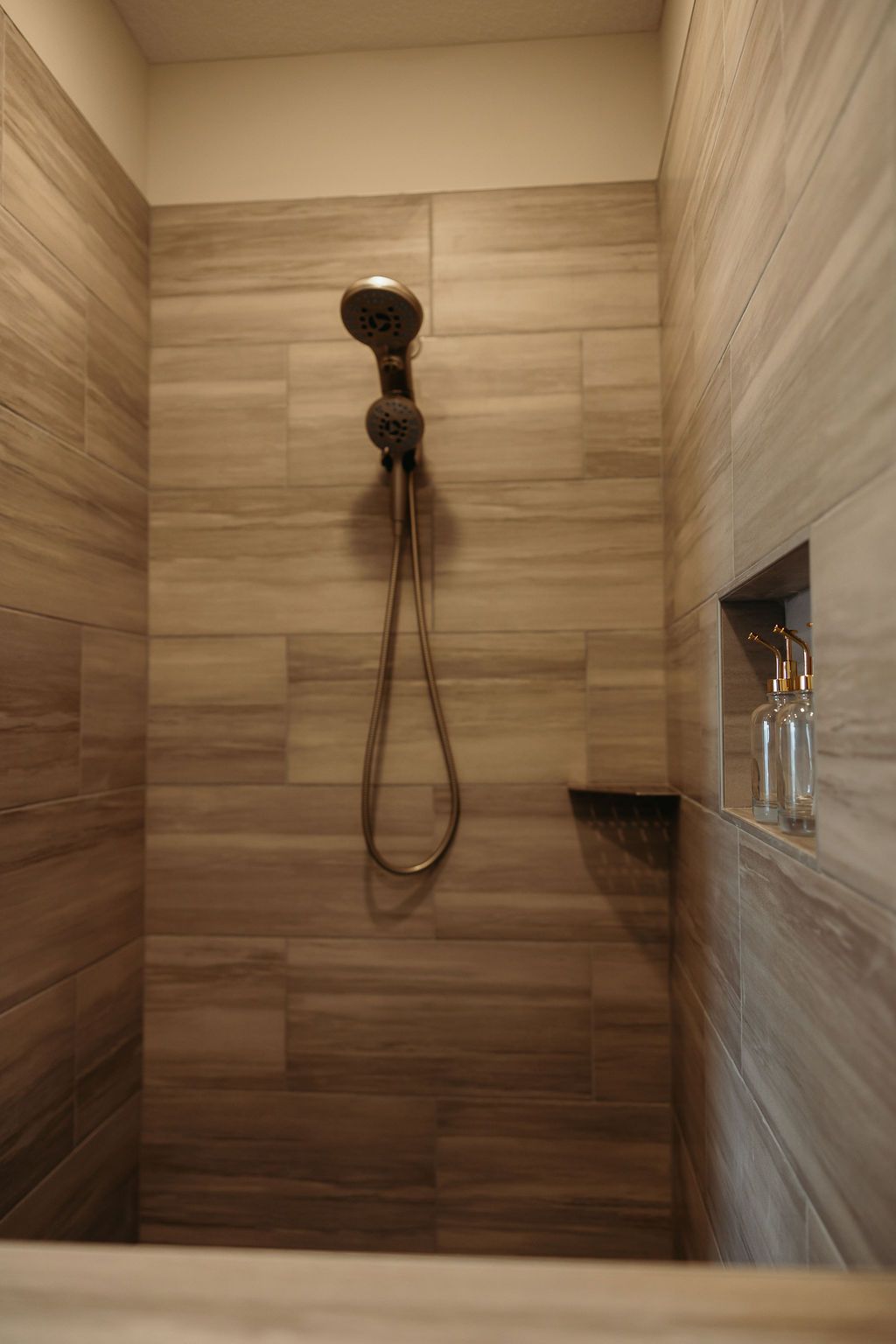 Shower with brown tile, a handheld showerhead, and a small built-in shelf with a soap bottle.