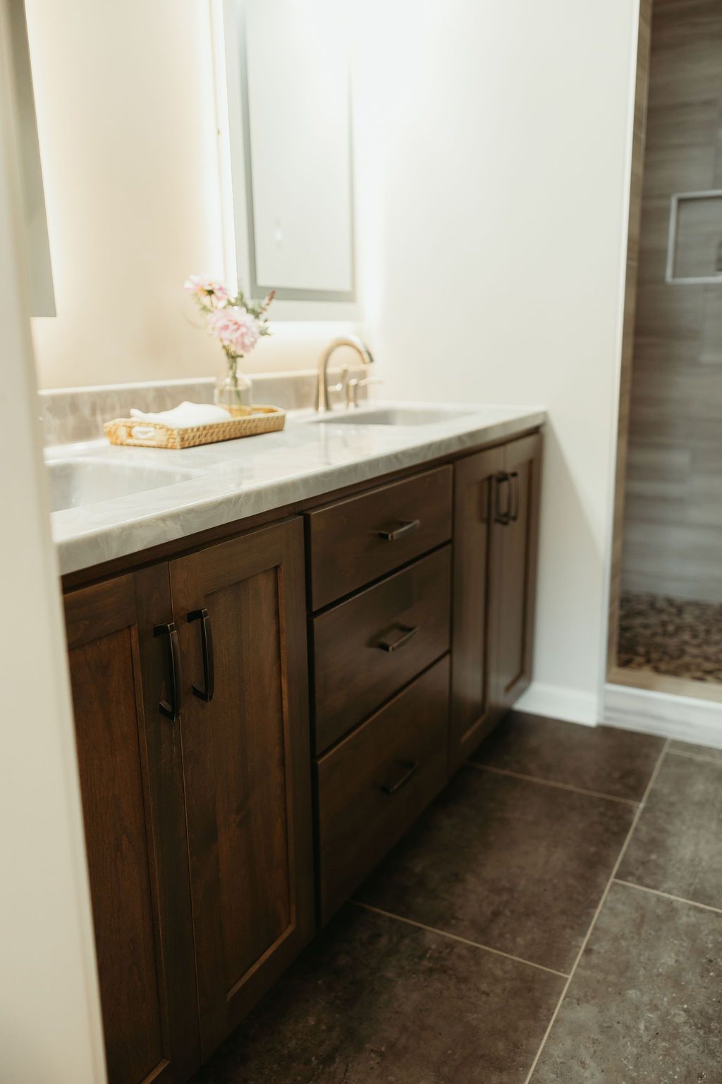Dark wood bathroom vanity with light countertop, sinks, and gold faucet. Tile floor, white walls.