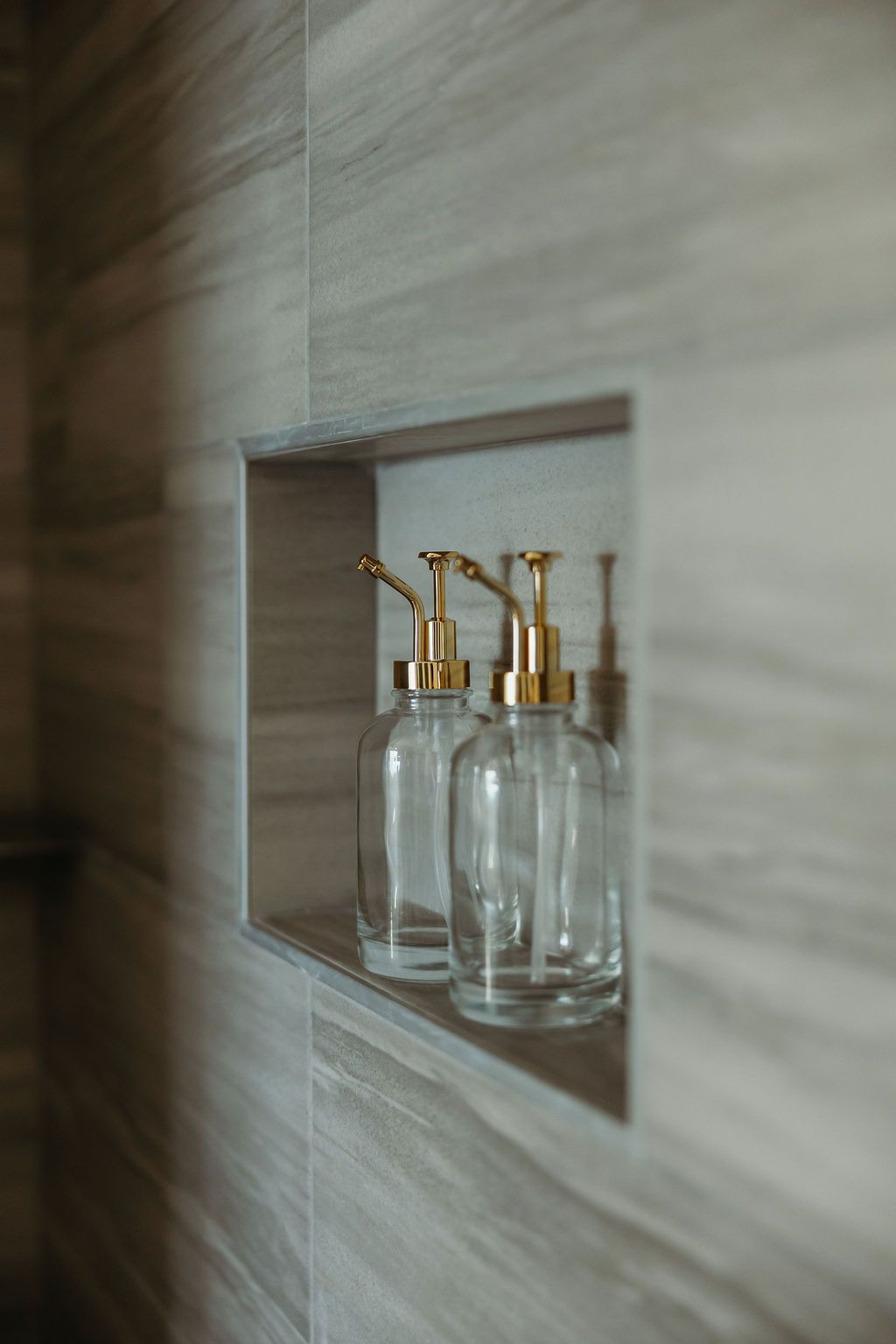 Two clear glass bottles with gold pumps in a recessed shower shelf.