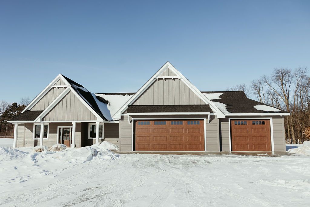 Gray house with three-car garage, brown garage doors, and snow-covered ground under a clear blue sky.