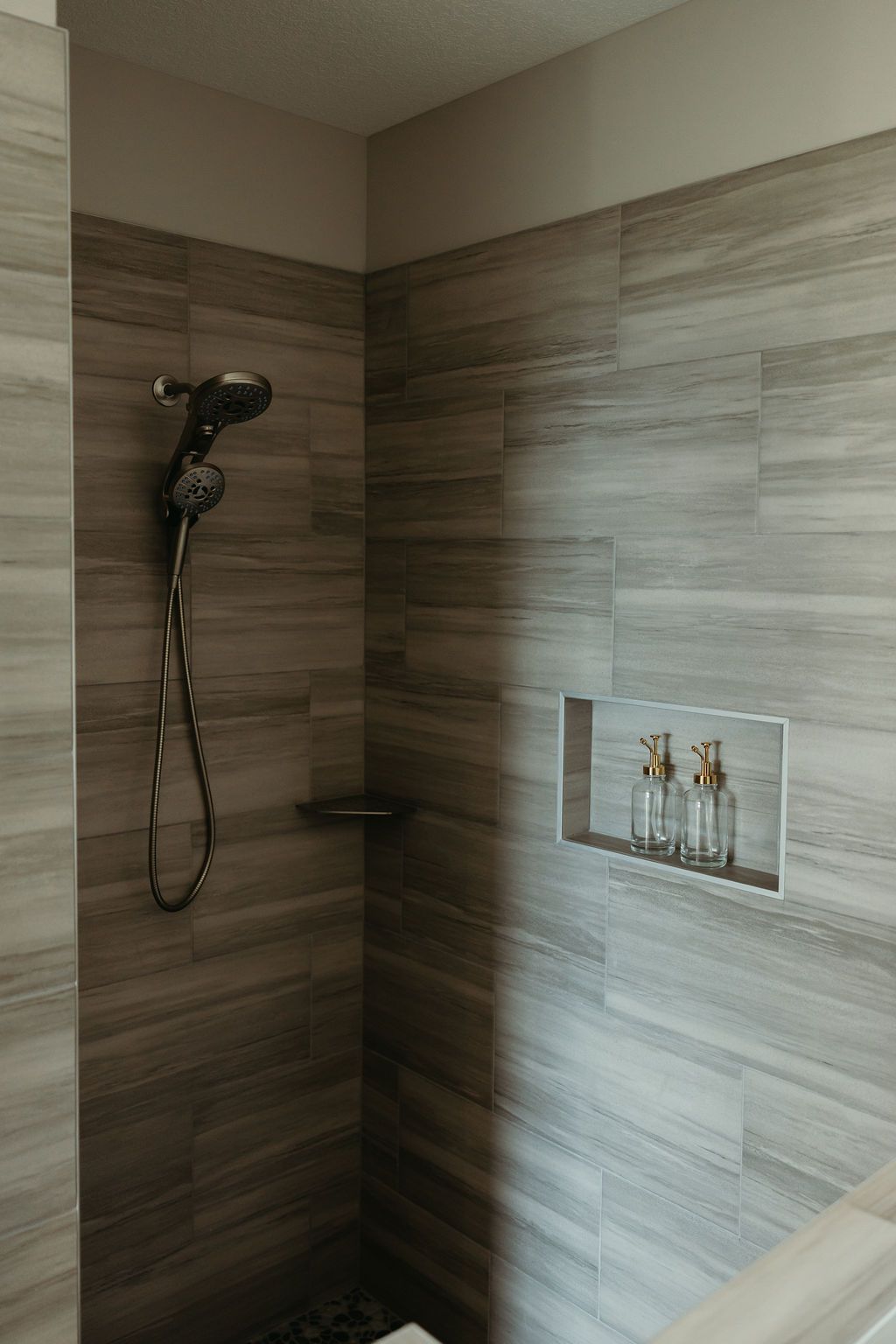 Shower with gray tiled walls, showerhead, built-in shelf, and recessed soap/shampoo niche.