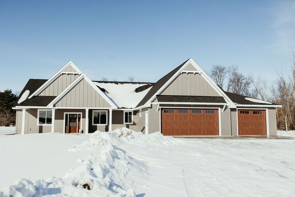 A snow-covered house with gray siding and brown garage doors on a sunny day.