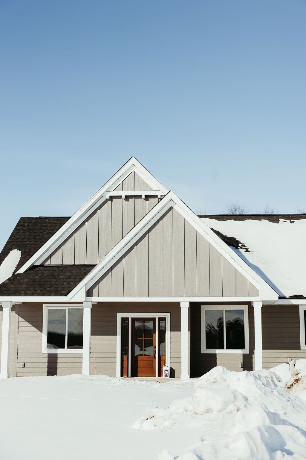A light gray house with a brown door, white trim, and a snow-covered yard on a sunny day.