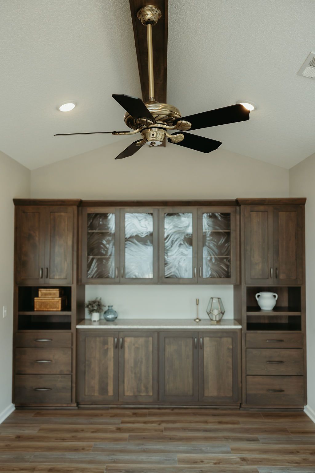 Dark wood built-in cabinetry with glass-fronted cabinets, a countertop, and a ceiling fan in a room with a sloped ceiling.