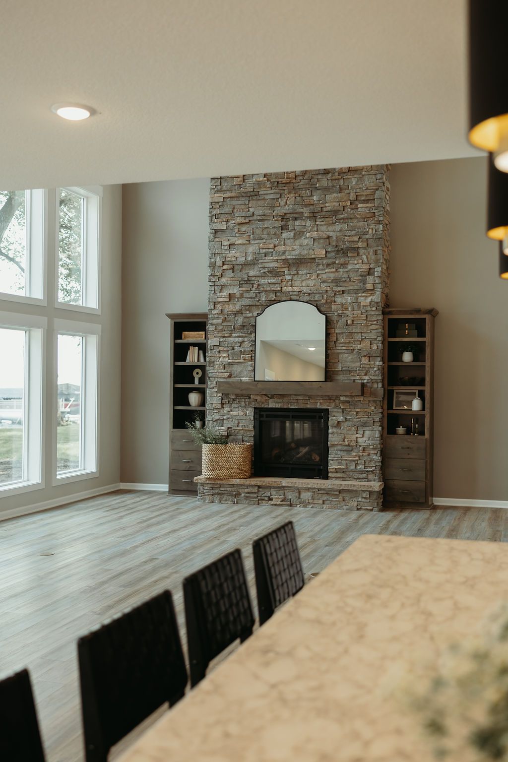 Living room with a stone fireplace, built-in shelving, large windows, and grey wood flooring.