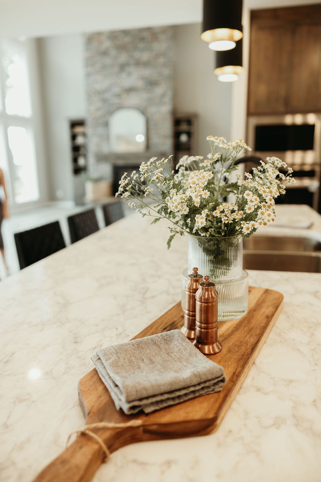 Wooden cutting board with flowers, salt/pepper shakers, and a folded cloth on a marble countertop in a kitchen.