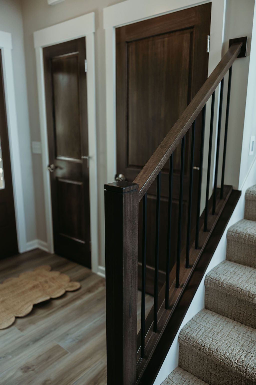 Dark wood doors, stairwell, and railing with black vertical spindles. Light wood-look flooring and carpeted stairs.
