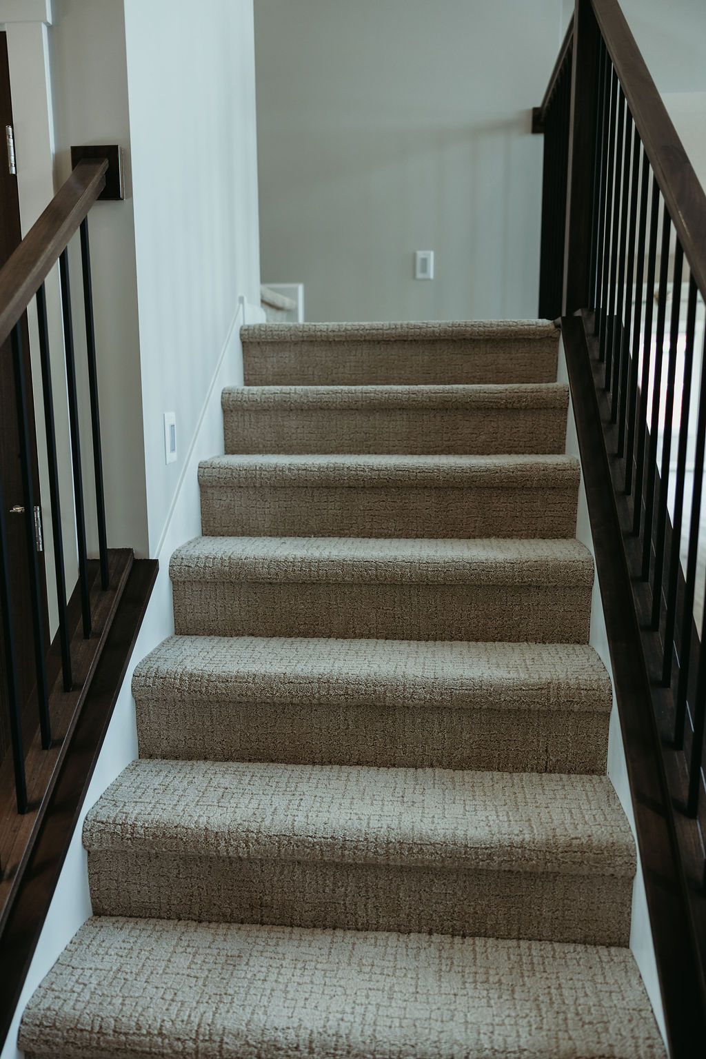 Staircase with carpeted steps and dark brown handrails, leading to a light gray wall.