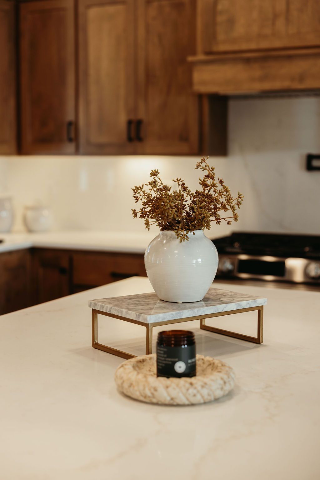 White vase with dried flowers on marble stand, candle, and stone bowl on kitchen countertop.