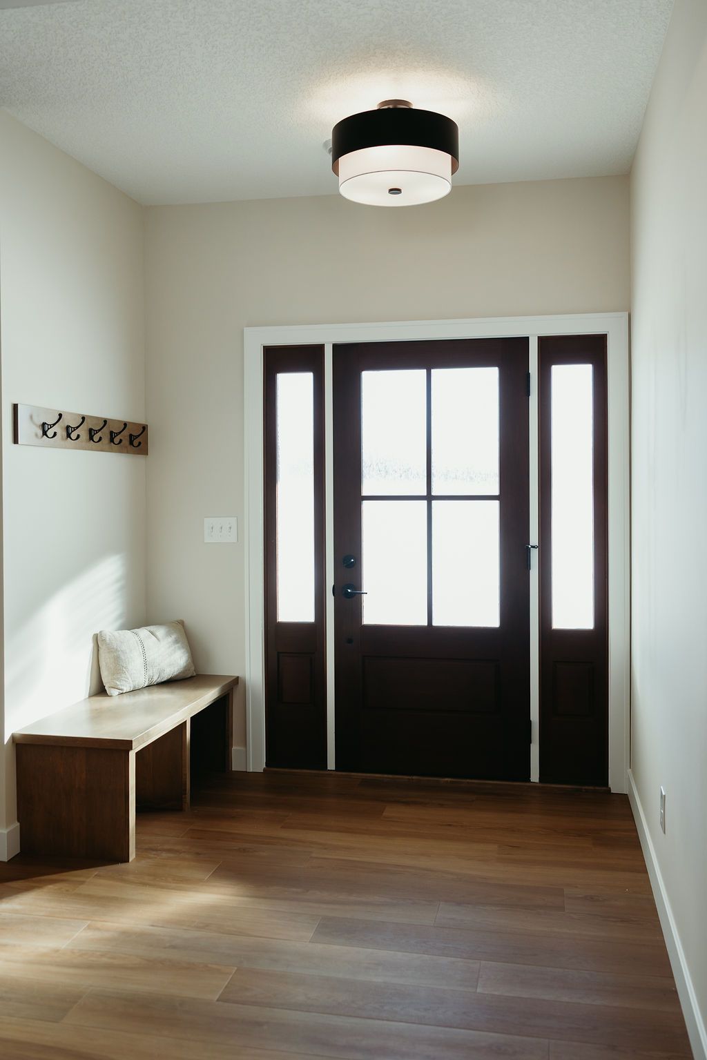 Entryway with dark brown door, bench, coat rack, and flush-mount light. Wooden floor and light walls.