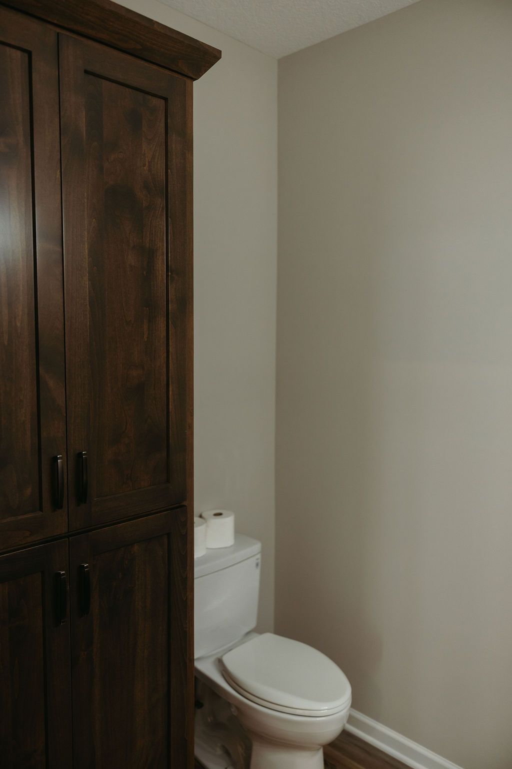 Tall, dark wood storage cabinet next to a white toilet in a bathroom with beige walls.