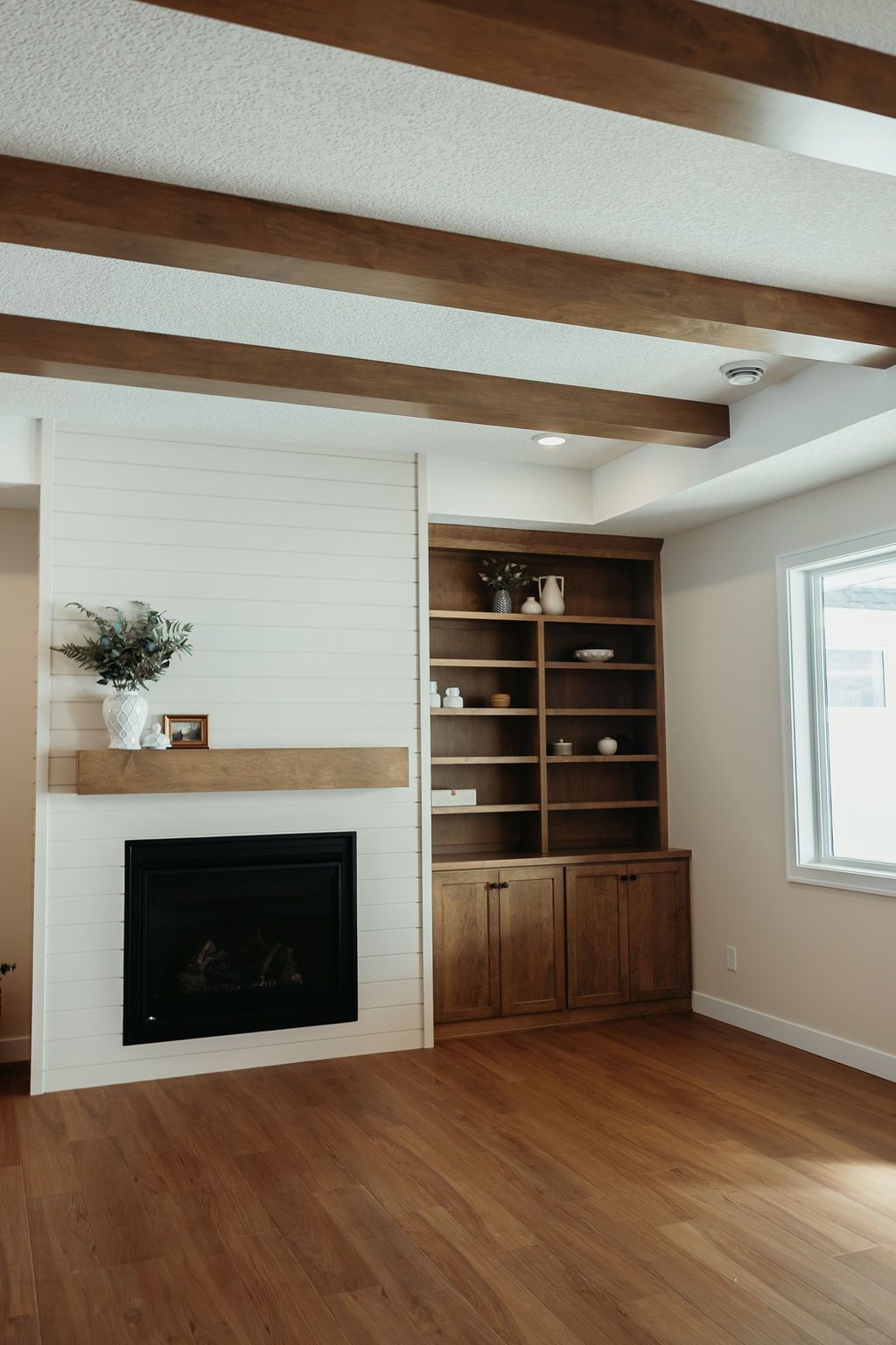 Living room with white brick fireplace, wooden built-in shelving, wood beams on ceiling, and wood flooring.
