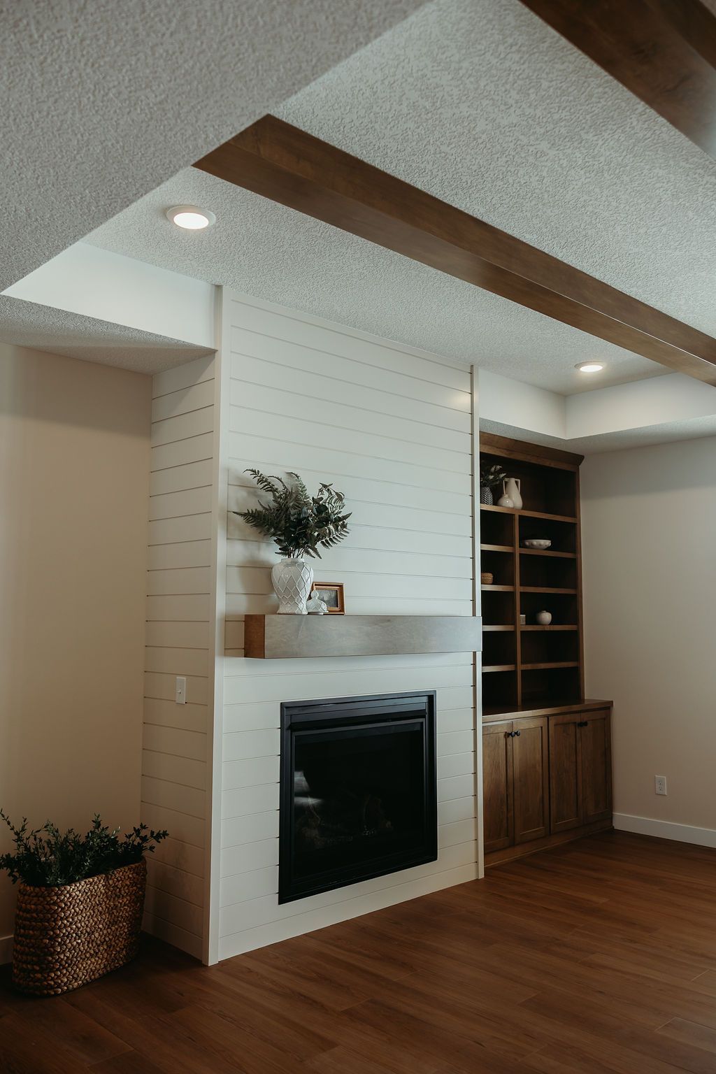 A fireplace with white shiplap, a gray mantel, and a dark, modern fireplace. Wooden beams and built-in shelving are visible.