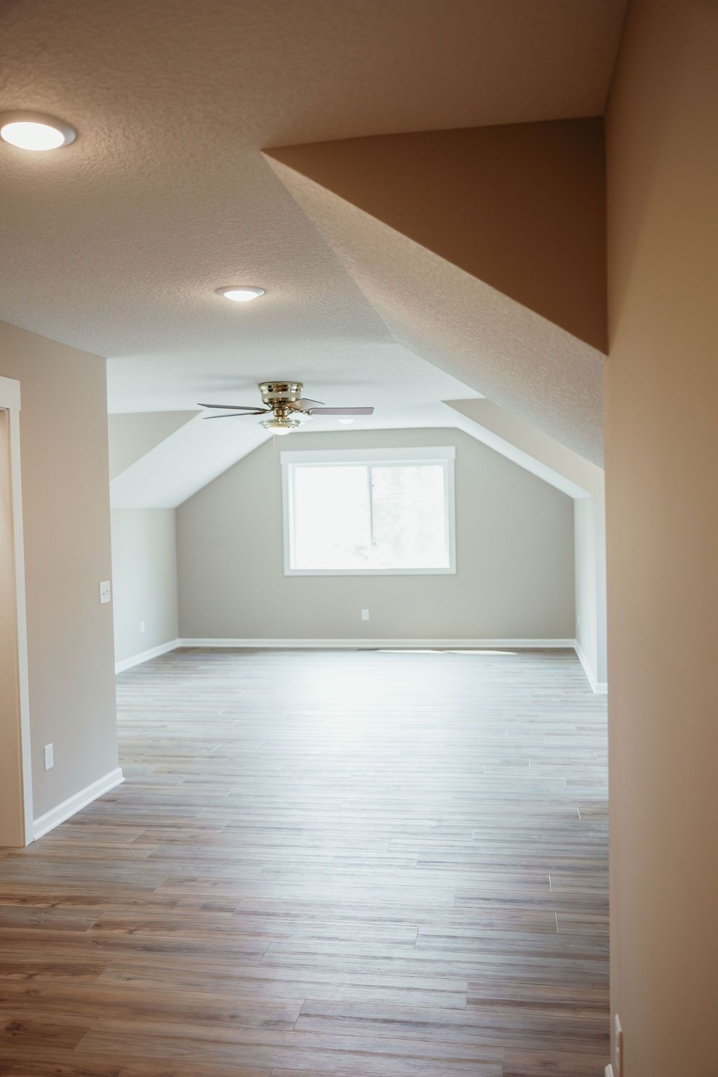 Empty room with angled ceiling, window, and wood-look flooring. Light gray walls, tan trim.