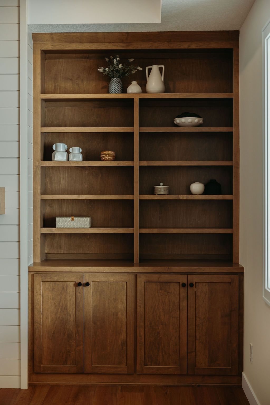 Wooden built-in bookshelf with multiple shelves and cabinet doors, filled with decorative items, against a white wall.