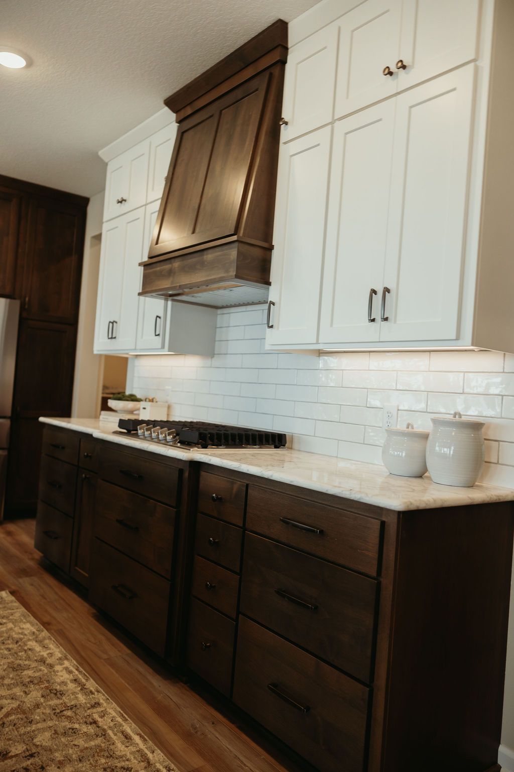 Kitchen with white upper cabinets and dark brown lower cabinets and range hood; white tiled backsplash.
