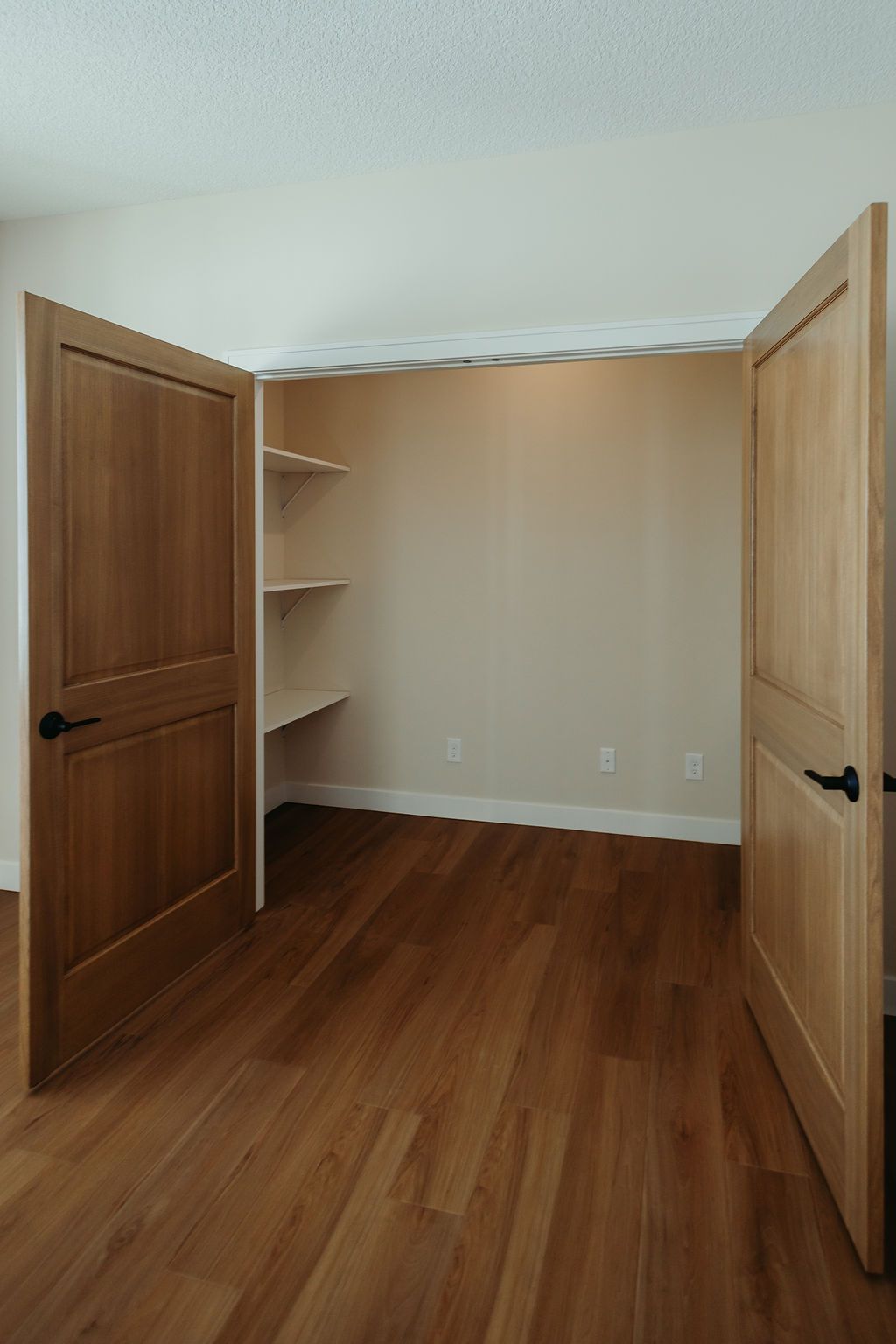 Closet with open wooden doors, shelves, and hardwood floor. The walls are neutral colors.