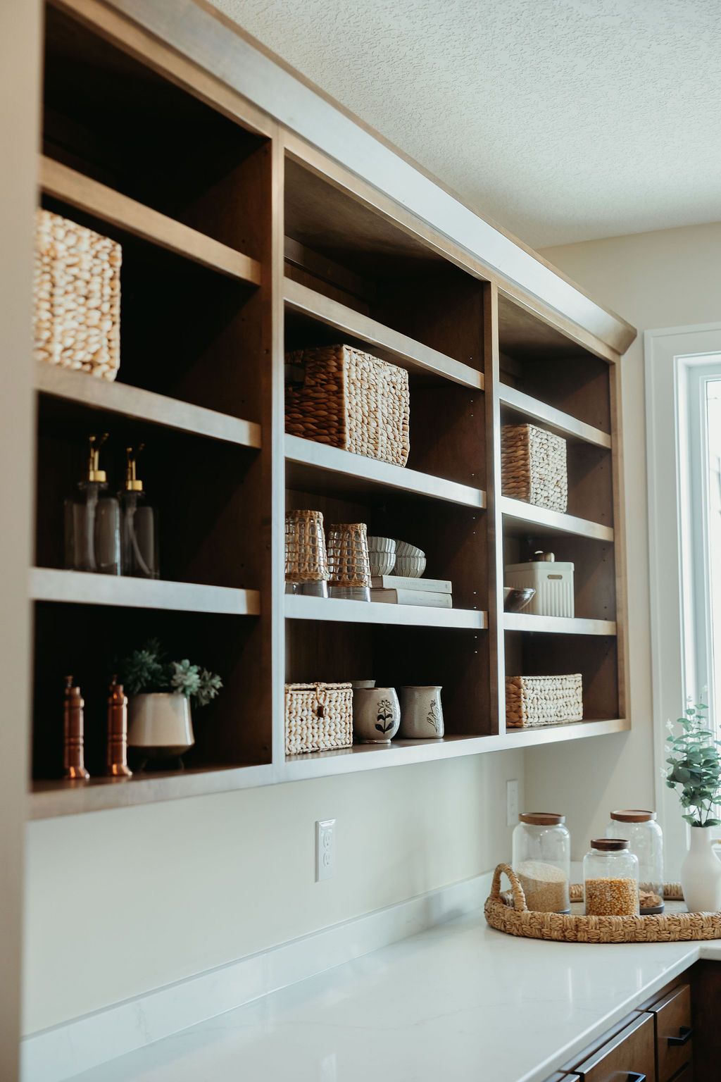 Built-in kitchen shelves with decorative items, including woven baskets, jars, and bottles, above a countertop.