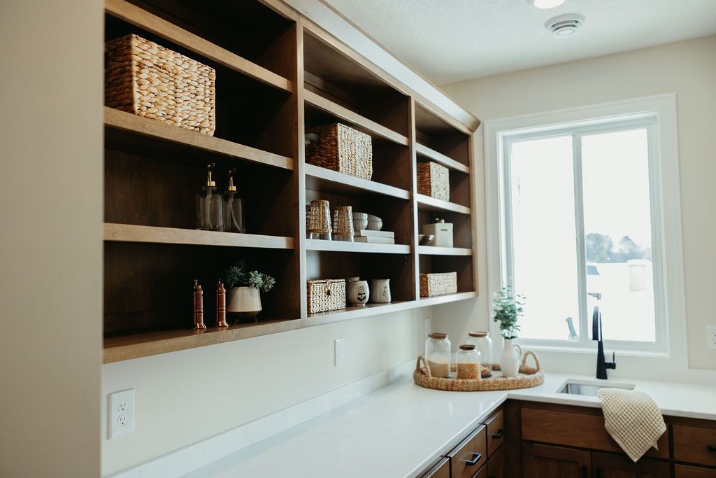 Built-in wooden pantry shelves with baskets and jars, next to a sink with a window, in a kitchen.