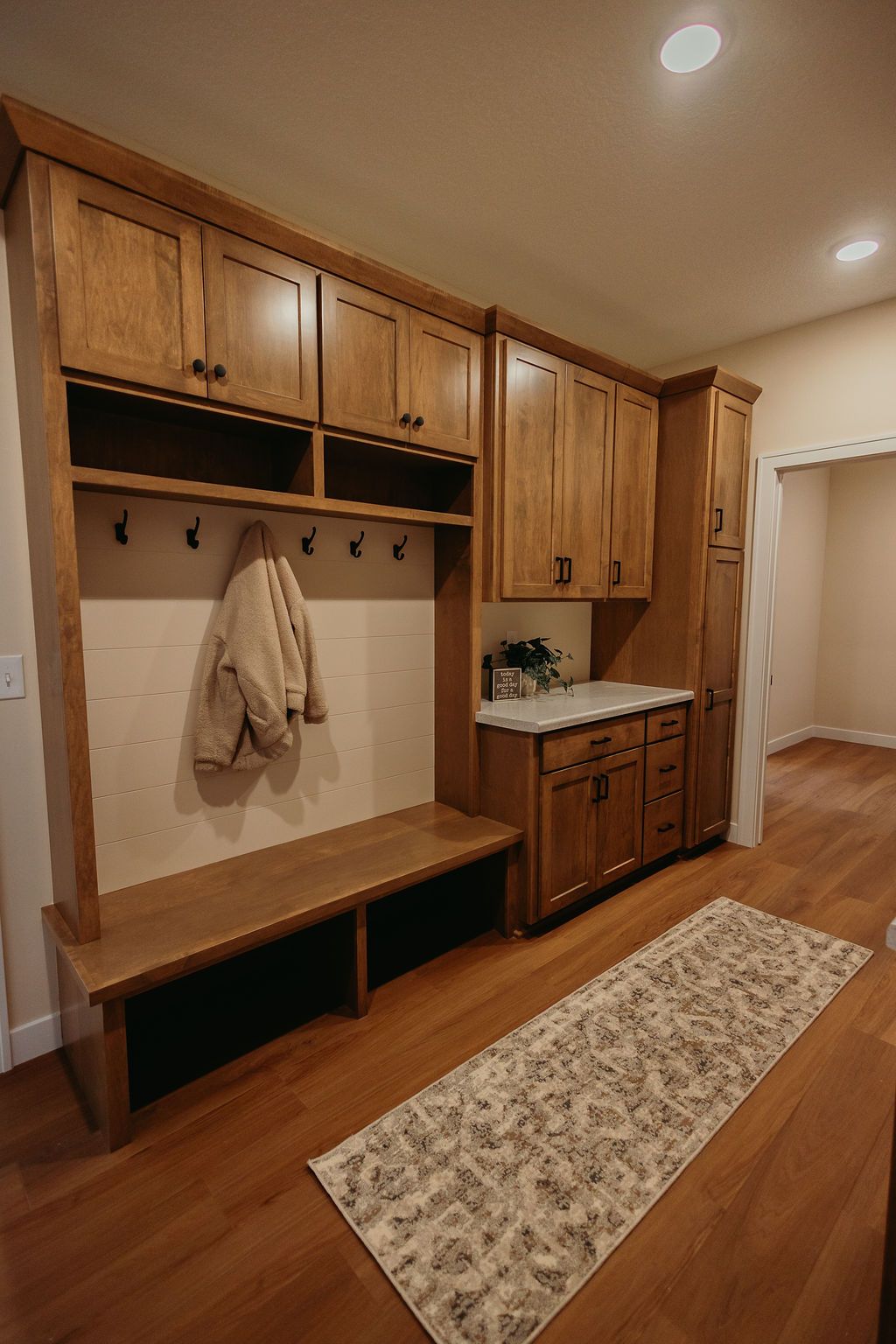 Wooden mudroom with built-in bench, cabinets, and hooks; tan jacket hangs.
