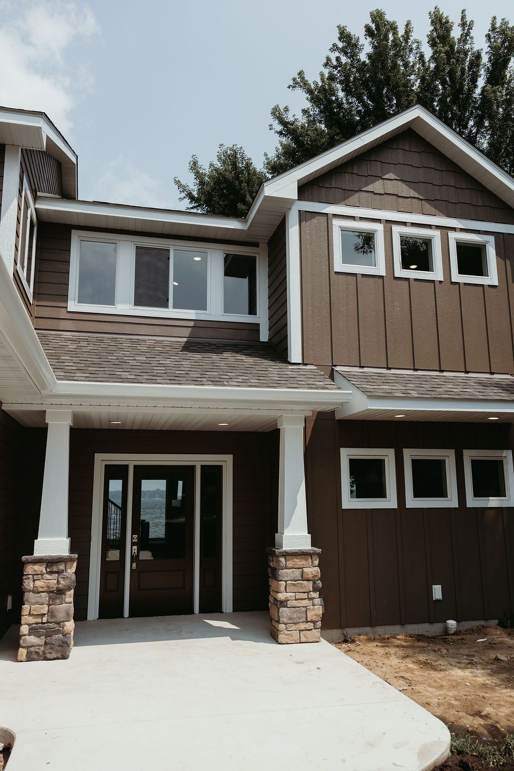 Brown two-story house with white trim. Entryway with stone pillars, windows, and a concrete path.