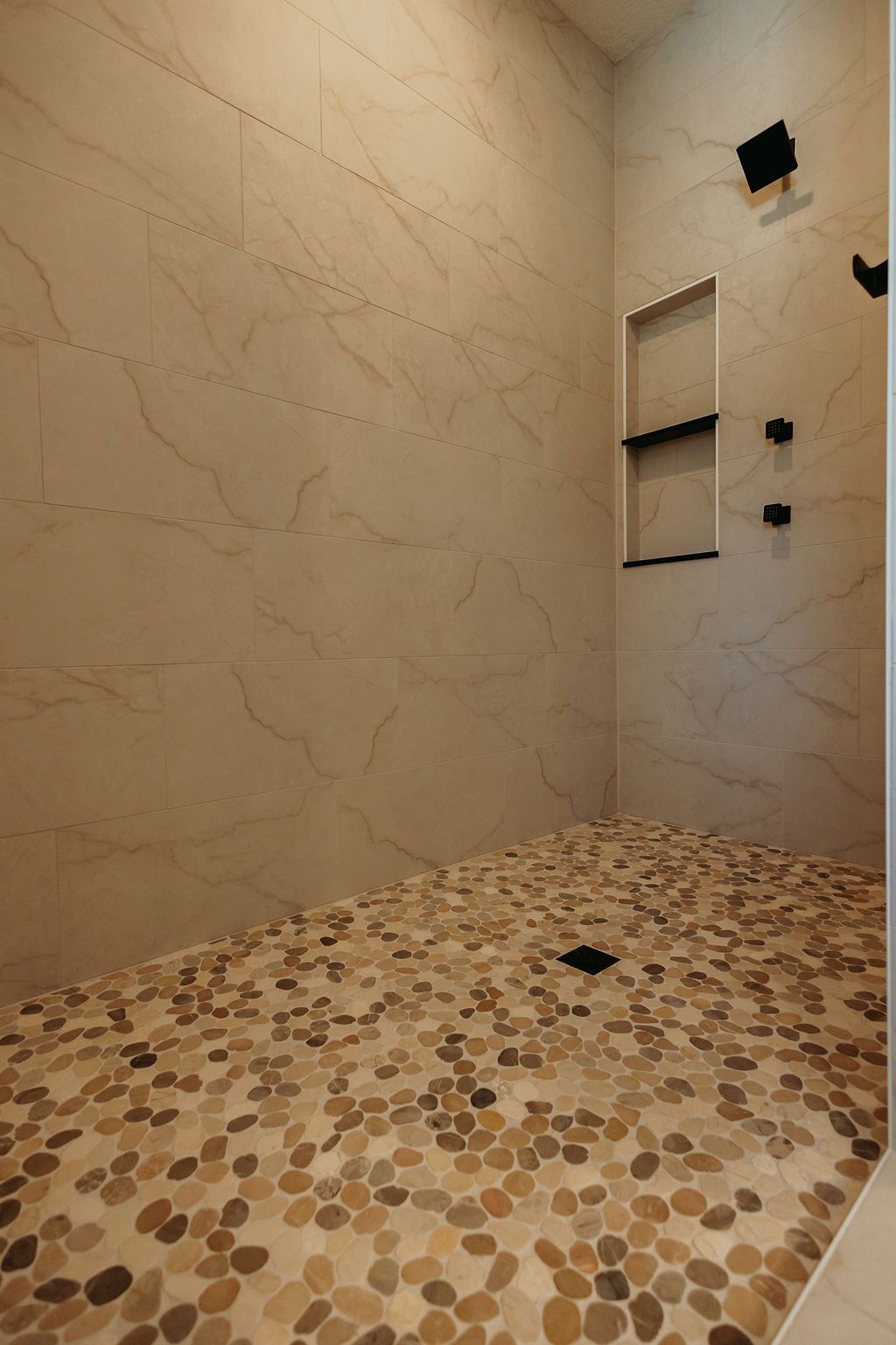 Shower with pebble floor, beige marble-like tile walls. Black shower fixtures and recessed shelf.