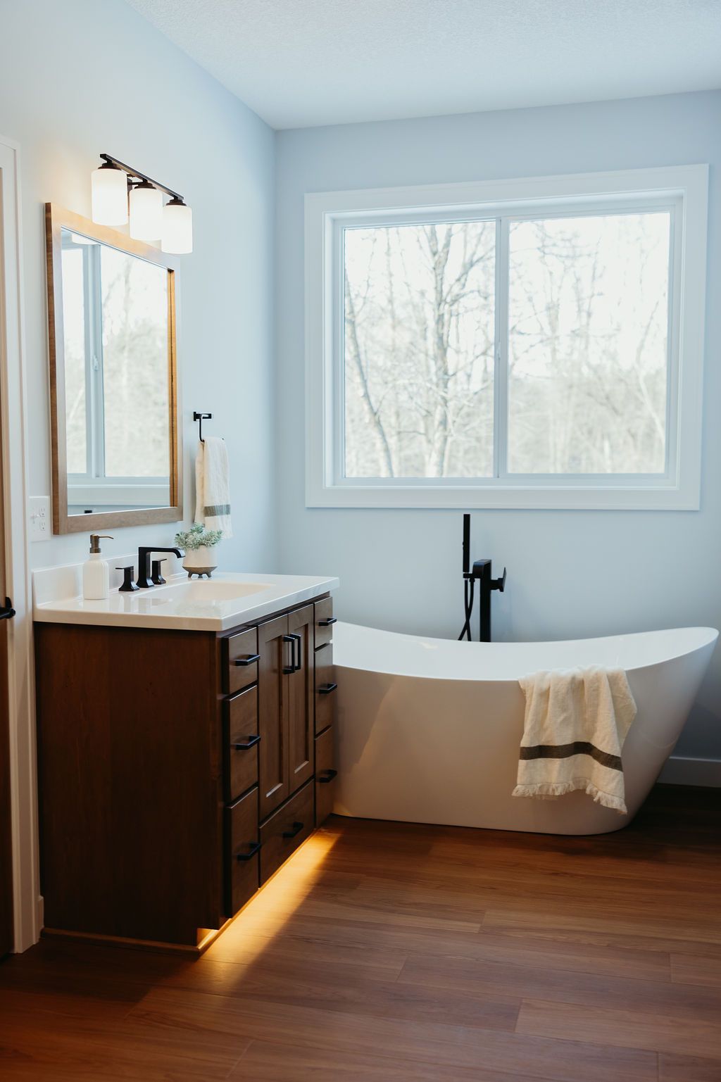 Bathroom with wooden vanity, white bathtub, large window, and light blue walls.