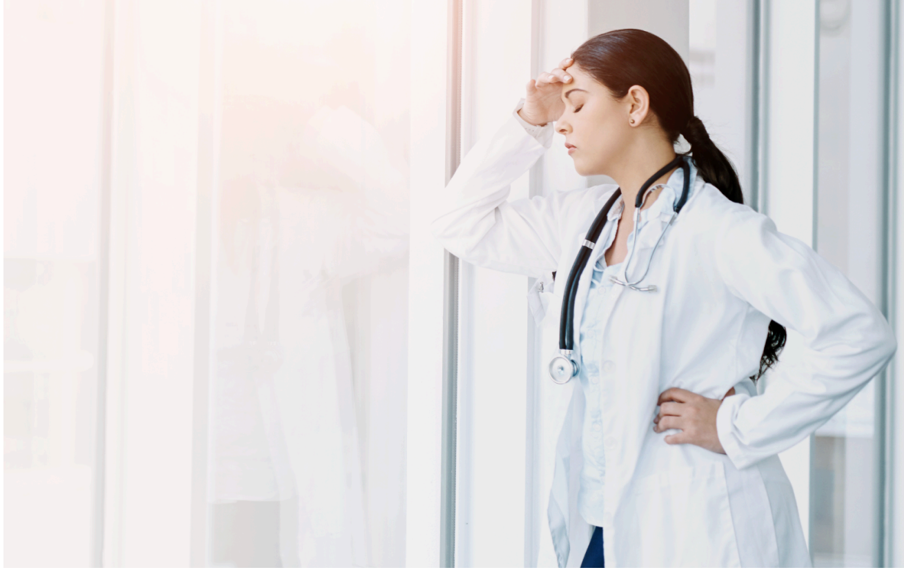 Doctor in white coat, hand on forehead, looks stressed by a window.
