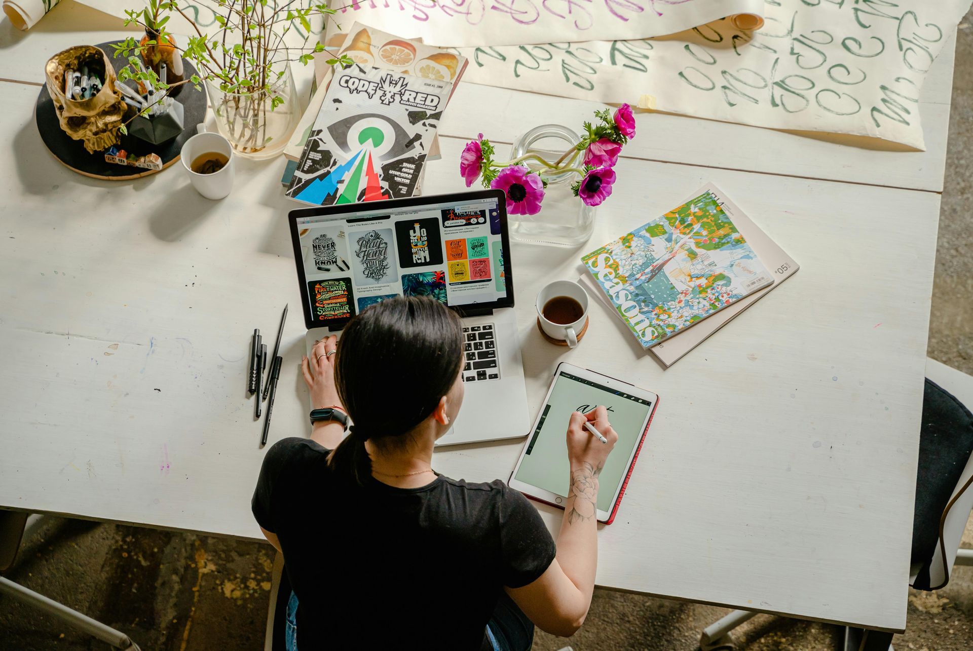 Person seated at white table, drawing on tablet, laptop open with artwork, coffee cups, and flowers.