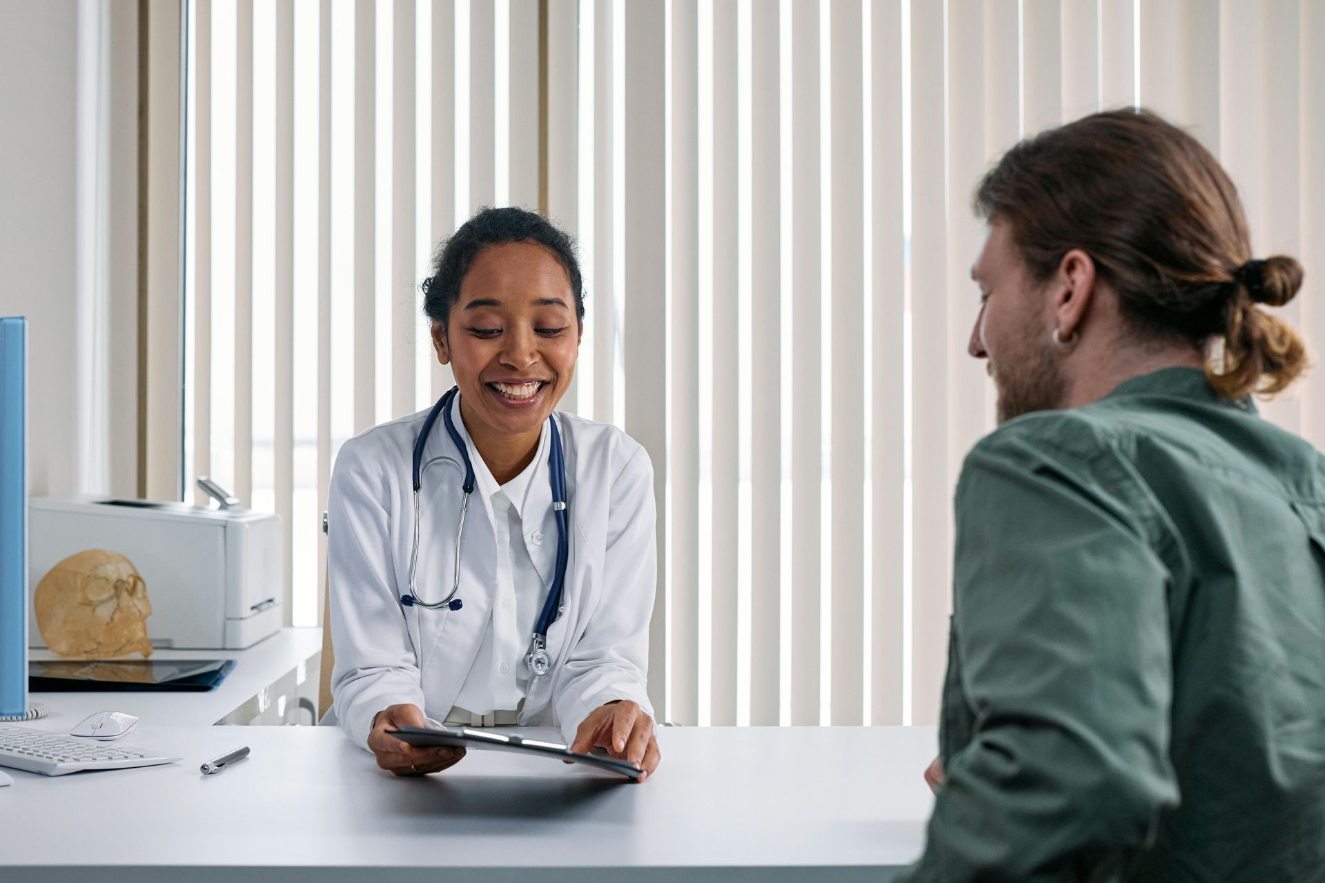 Doctor smiling, talking to patient in office. Woman doctor wears lab coat. Man sits opposite her.