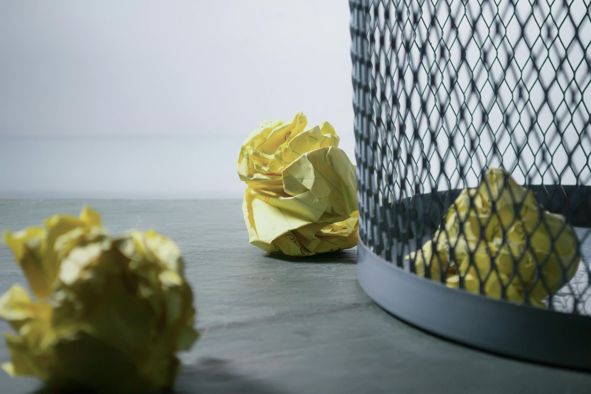 Yellow crumpled papers near a black wire trash can on a gray surface.