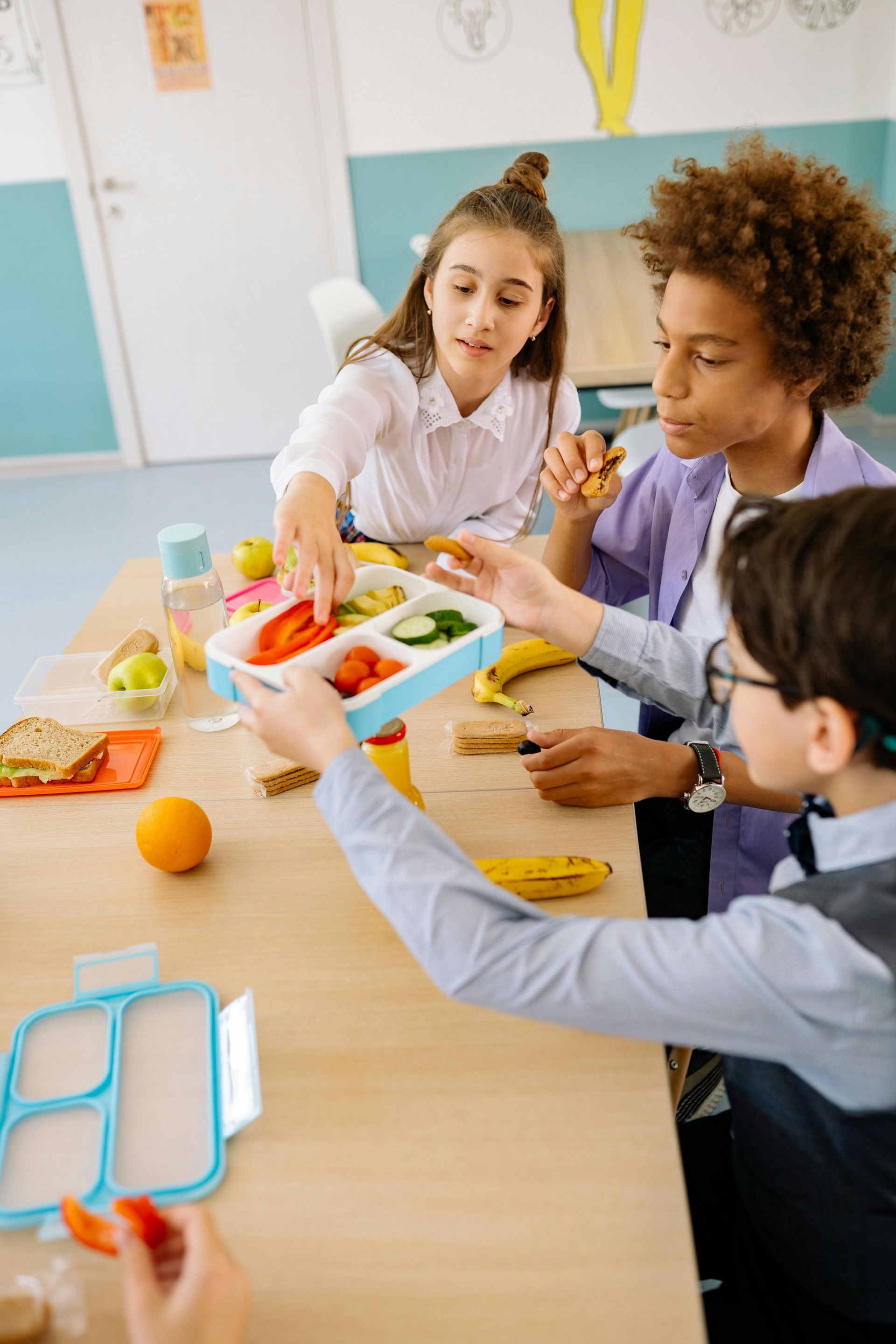 Three students sharing a lunchbox at a table, one reaching for a tomato slice.
