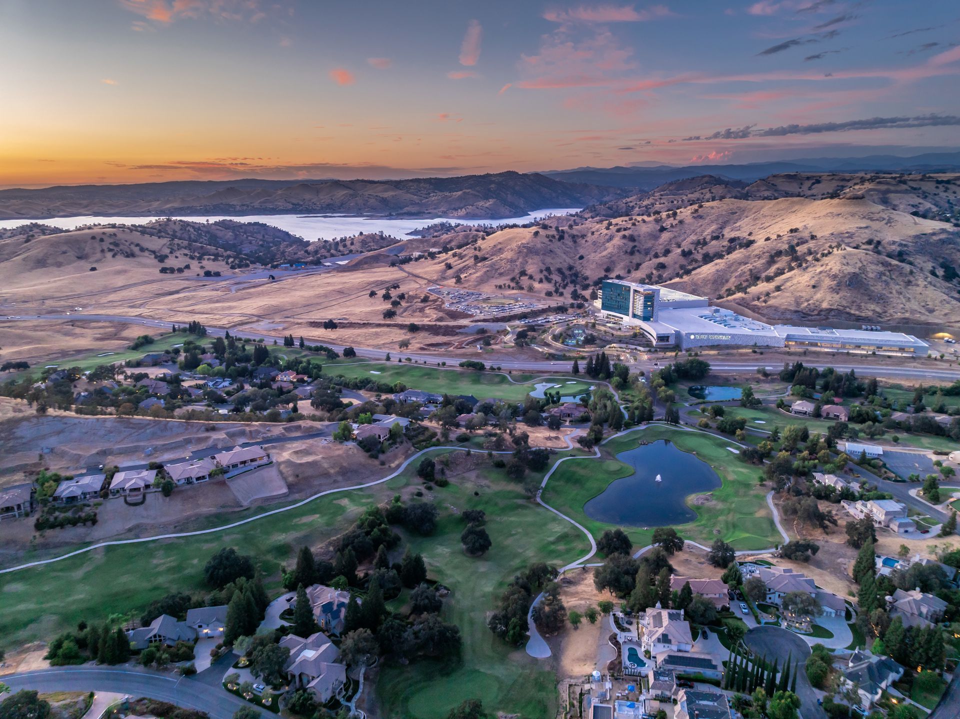 Aerial view of rolling hills with golf course, houses, and a large body of water in the background at sunset.