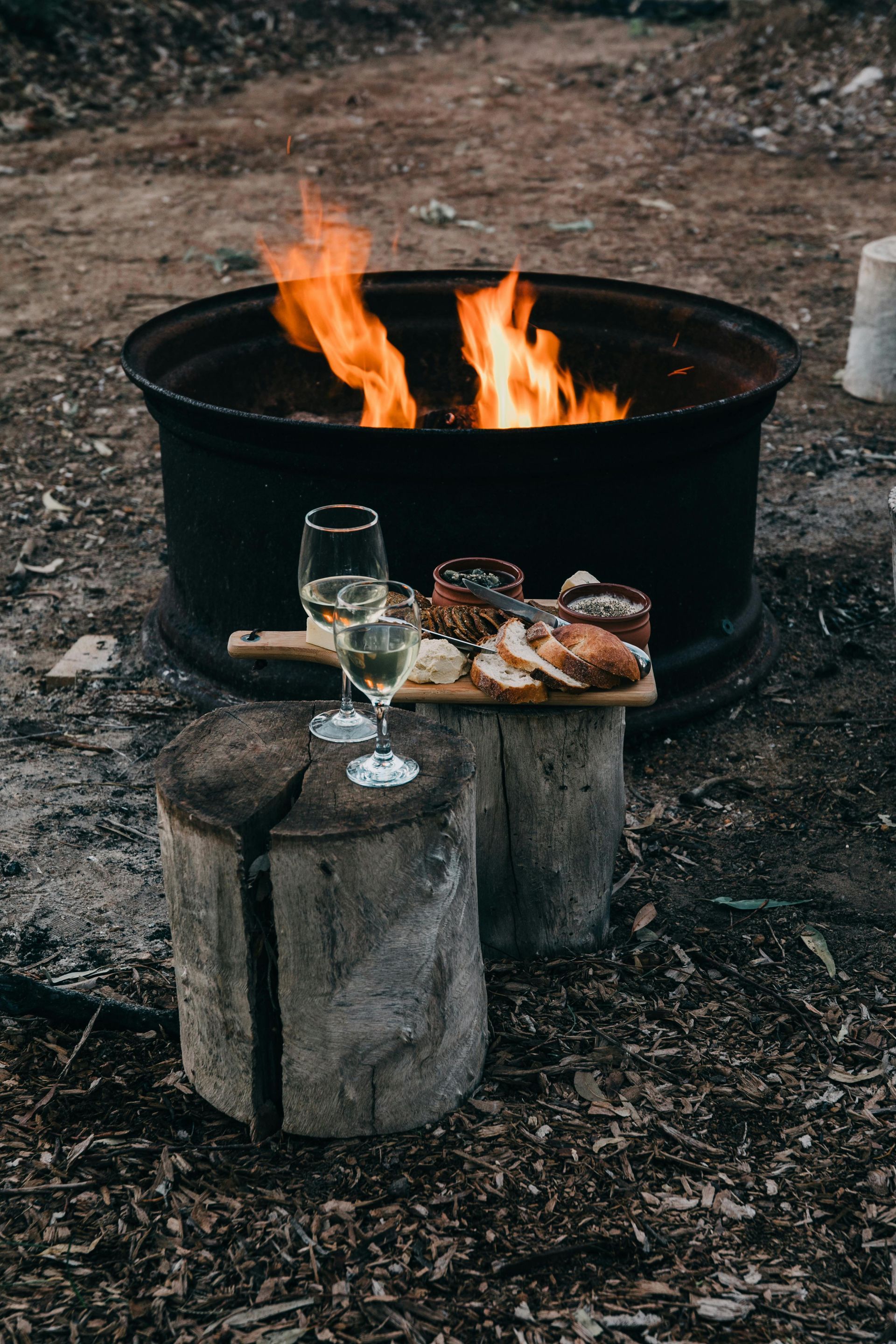 Fire pit with flames, food and wine on wooden stumps in an outdoor setting.