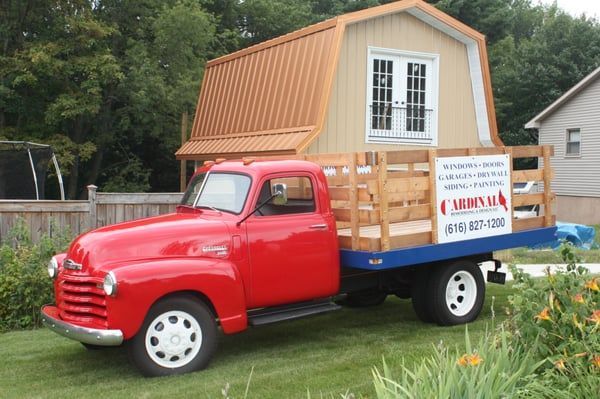 Red vintage truck carrying a small tan shed with a copper roof on a green lawn.