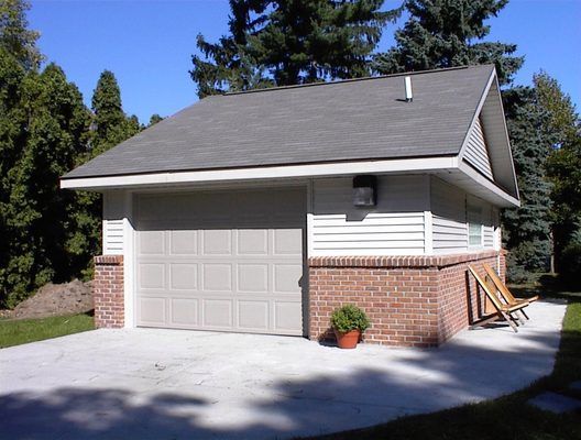 Garage with gray door and roof, brick base, white siding, concrete driveway, and outdoor chair.