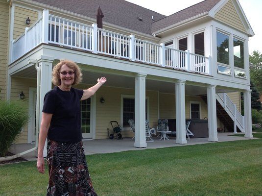 Woman standing in backyard, pointing towards a two-story deck with white railings and a yellow house.