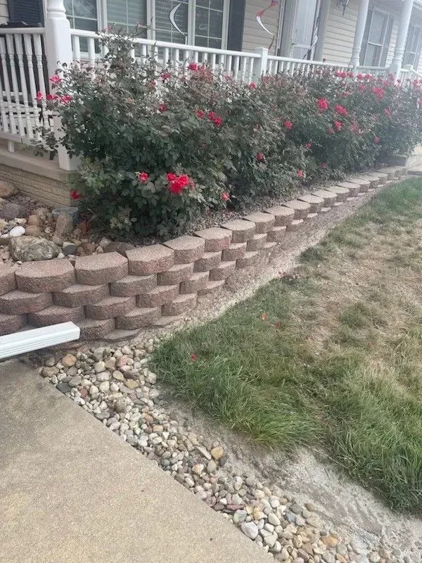 A low retaining wall made of reddish-brown blocks borders a flower bed filled with red rose bushes and small gray rocks.
